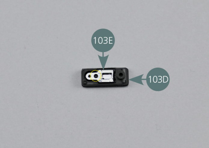 Dispón el soporte de manilla de puerta (103D) con la parte interior hacia arriba como indica la imagen. Introduce la manilla de puerta (103E) en el soporte, asegurándote de que el pin esté correctamente alineado con el orificio (círculo amarillo). La manilla de puerta es así montada por el reverso.