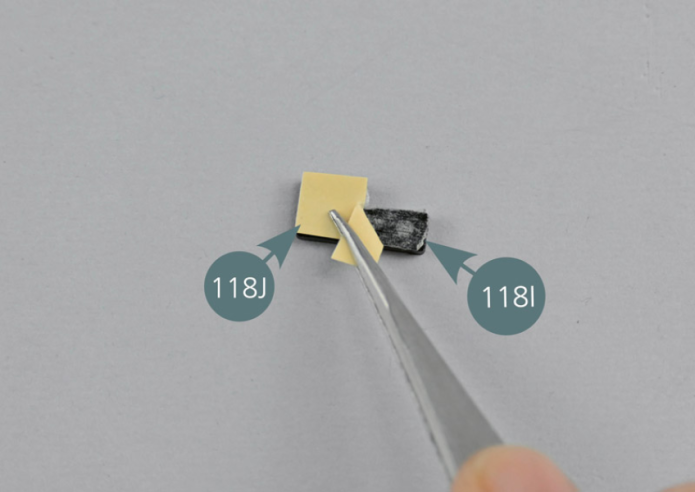 Remove the protective film from a double-sided adhesive tape (118J). Apply the adhesive (118J) onto one of the hinge spacers (118I).Remove the second protective film from the adhesive tape (118J) and attach the spacer (118I) to the left hinge (118D) as shown.