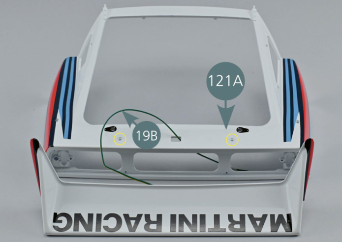 Passer le fil 19B dans le trou rectangulaire central du capot arrière 121A, de l’extérieur vers l’intérieur.Positionner l’aileron arrière 123A sur le capot arrière 121A en faisant coïncider les points de fixation avec les trous correspondants (cercles jaunes).Retourner l’ensemble et fixer l’aileron avec quatre vis AM.