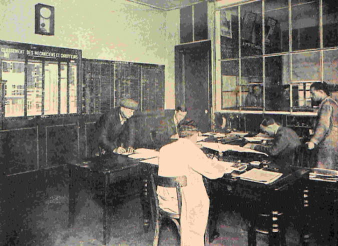 Le bureau de « la feuille », centre névralgique du dépôt, dans les années 1950. Les mécaniciens attendent leur feuille et les instructions verbales en restant dans le couloir, derrière le guichet vitré à droite. Aux murs : les tableaux et la craie pour la préparation des « feuilles de route ».© IXO Collections SAS - Tous droits réservés. Crédits photo © Collection Trainsconsultant-Lamming